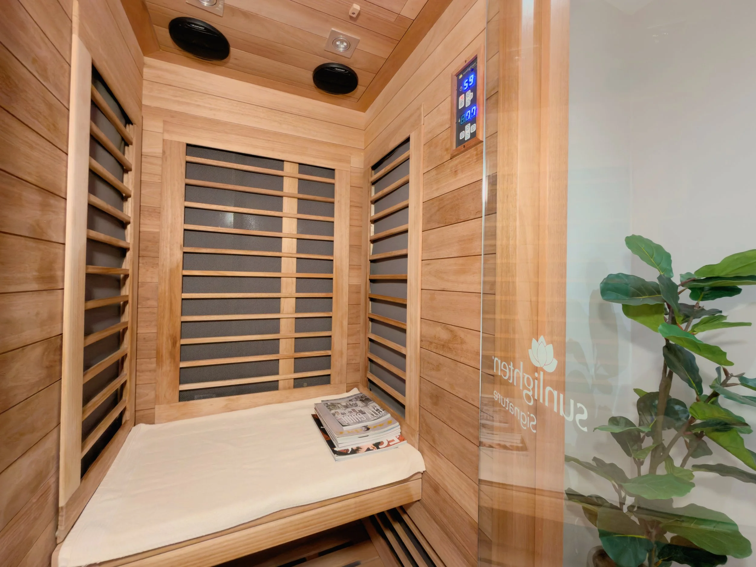 A small, wooden infrared sauna with a bench inside, a stack of magazines on the bench, and a glass door with a logo and a green plant next to it.