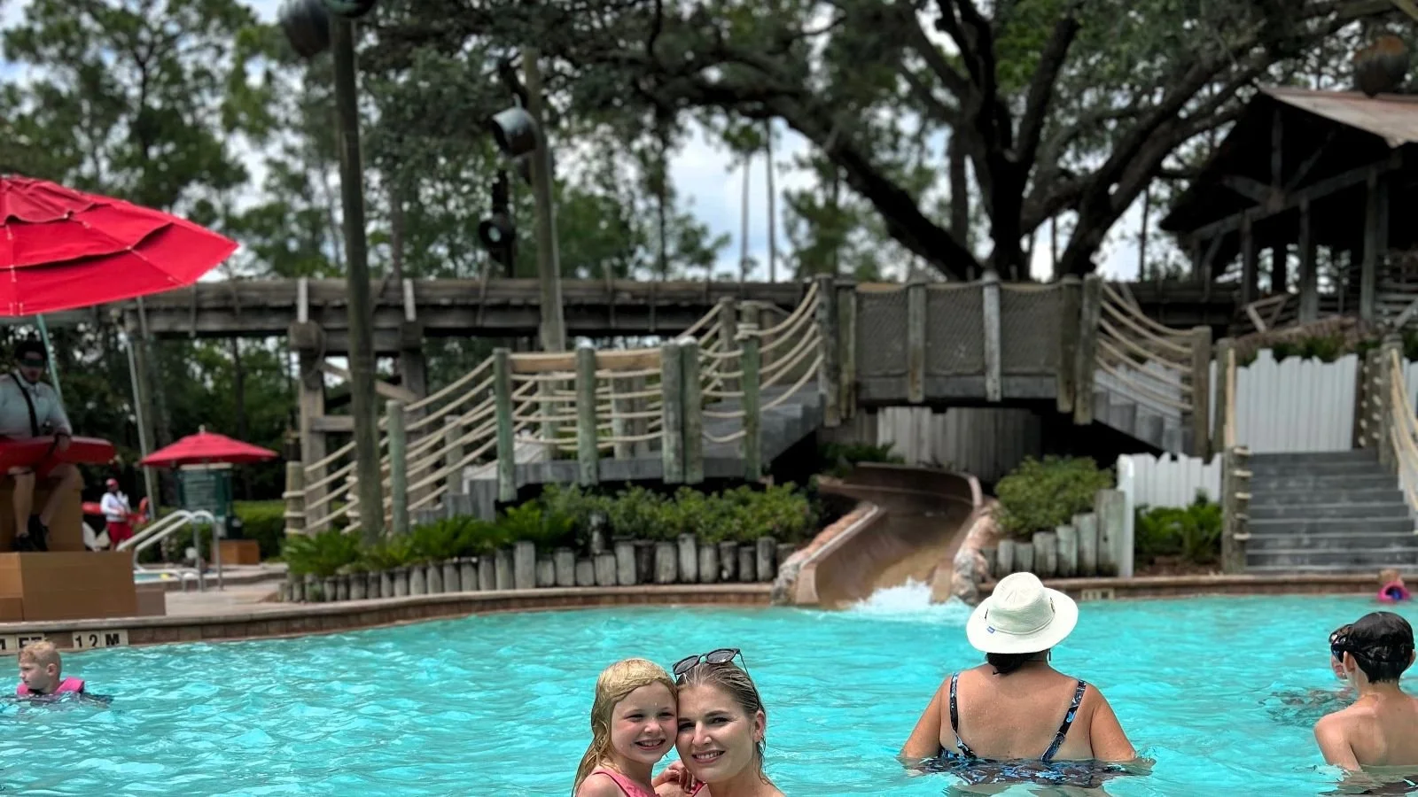 A mom and daughter pose in the foreground of the Ol' Man Island pool. Behind them is a water slide. It's at Port Orleans Riverside, a moderate disney world resort.