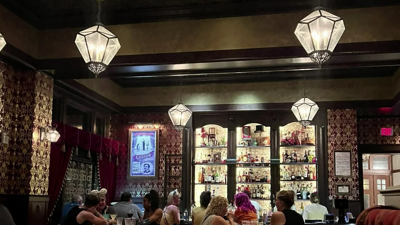 The interior of AbracadaBar, a bar in Boardwalk at Disney World. You can see an early 1900s magician aesthetic with dark wood, high contrast patterned wall paper, fancy chandeliers, and an extremely tall, backlit bar shelf. There are about 12 patrons