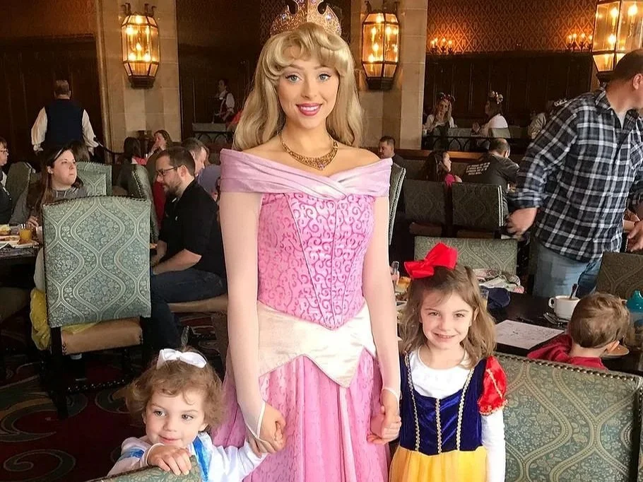 Two small girls in princess dresses hold hands and pose with Sleeping Beauty in Cinderella's Royal Table, a character dining experience in the Magic Kingdom castle. There are several guests eating behind them.