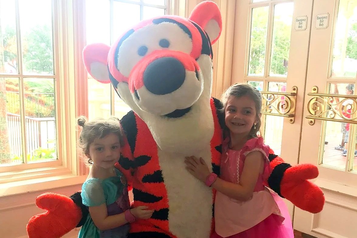 A photo of two girls in princess dresses with Tigger at The Crystal Palace, a character meal at Disney World. They are smiling and standing in front of a window, making for a poorly lit picture.