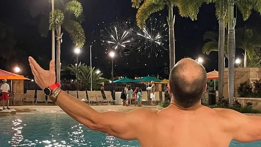 a man lifts his arms in mock wonder as he watches the fireworks at the Caribbean Beach Resort pool, a moderate resort at Disney World.