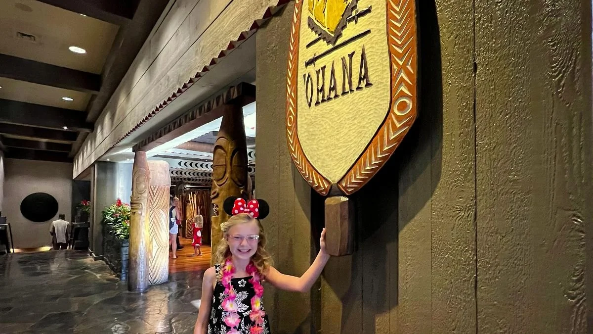 A girl smiles in front of the 'Ohana sign at the Polynesian Resort. You can see dark stained wood and intricately carved tiki columns. The floor is polished volcanic rock. 'Ohana has a Disney World character breakfast.