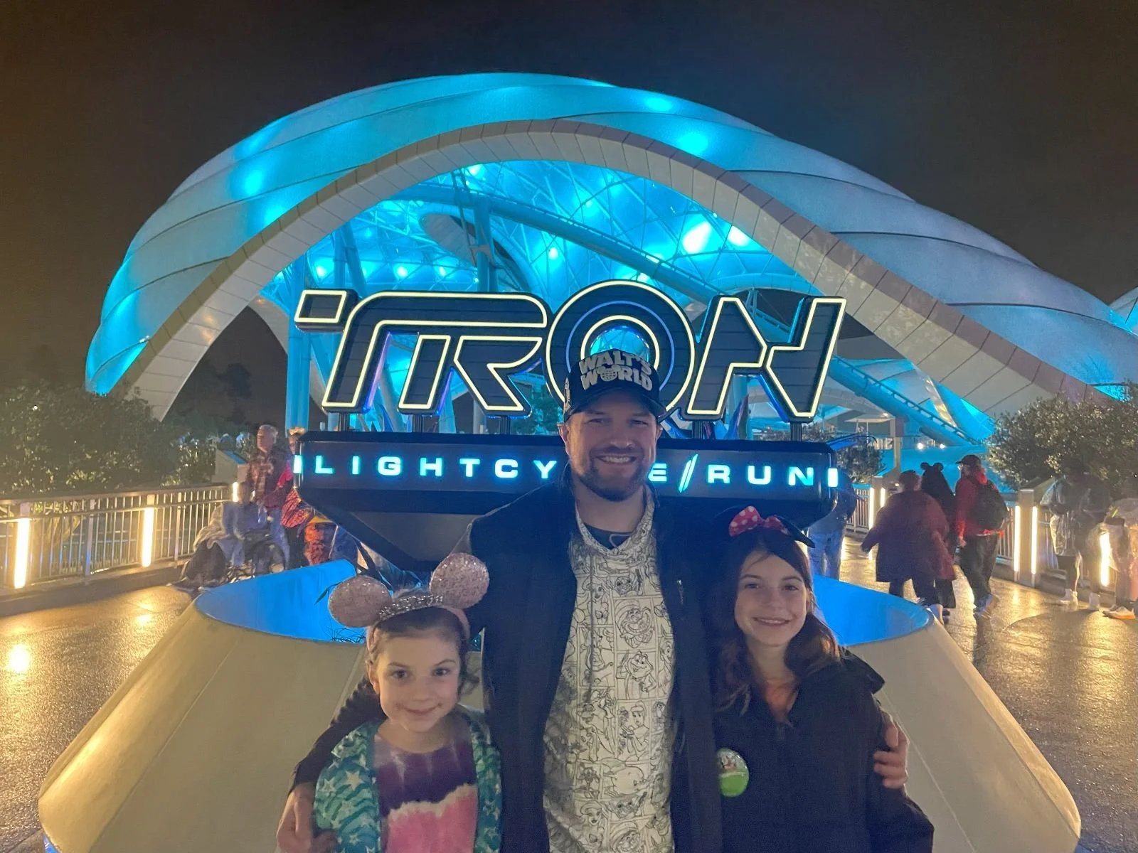 A dad and two daughters pose in front of the sign for Tron:lightcycle, a roller coaster in Magic Kingdom at Disney World. The dome above the ride is lit up with blue lighting.
