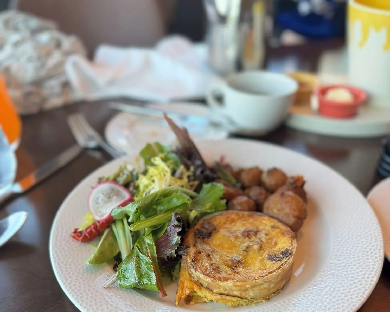 A photo of the Topolino's character breakfast quiche gruyere. It's a round, fist-sized quiche with salad and potatoes. Behind it you can see the paint bucket pastry bucket and the press pot.