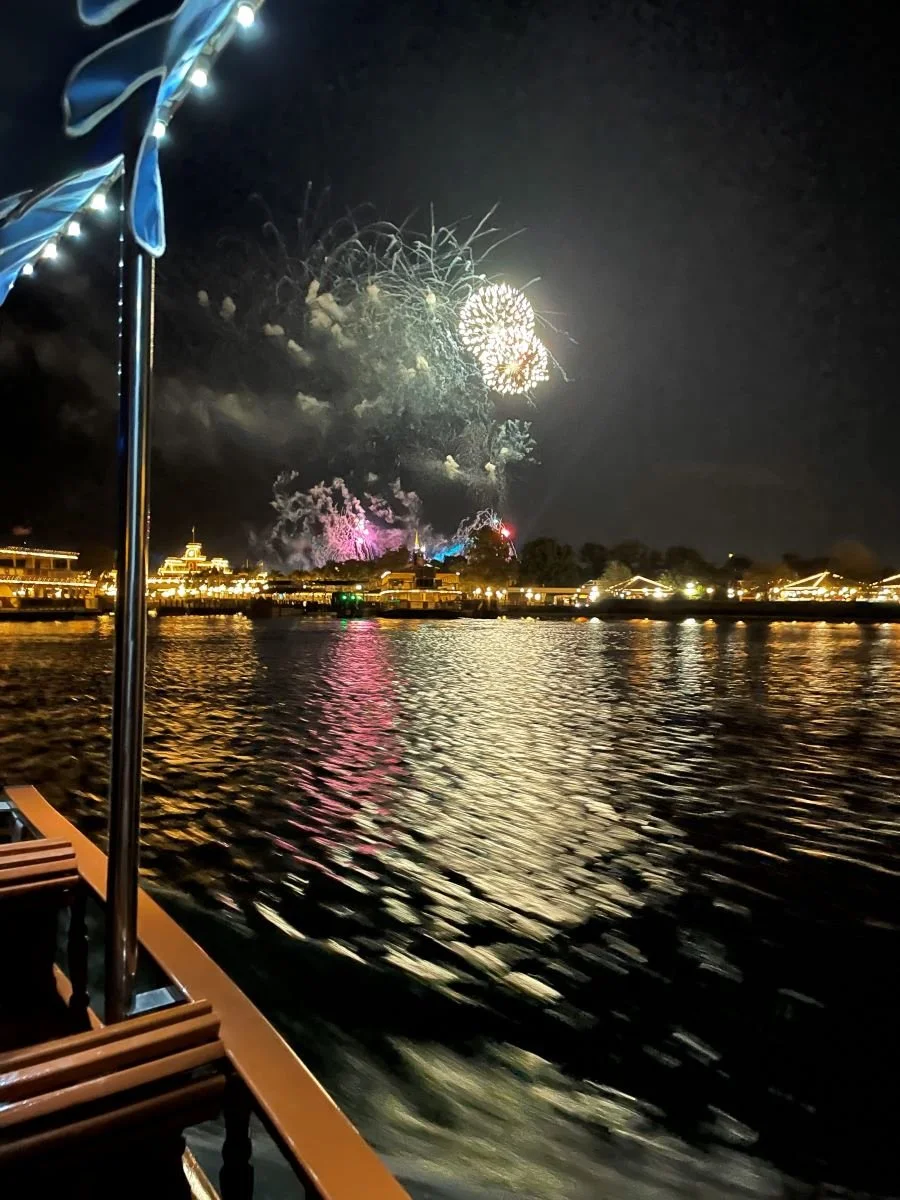 A photo from a small boat looking at fireworks over the entrance of Magic Kingdom at Disney World. The boat came from Wilderness lodge. You can see the lake and lots of lit up buildings beneath the fireworks.
