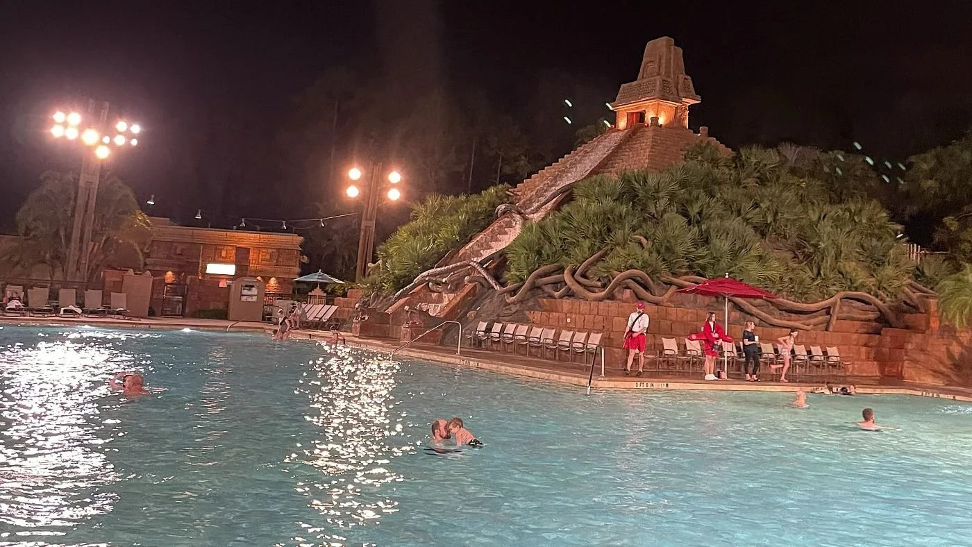 An image of the Lost City of Cibola pool at Coronado Springs in Disney World. A dad plays with a toddler in the foreground. There's enormous ziggurat covered in vines and plants, with a waterfall coming down the stairs.