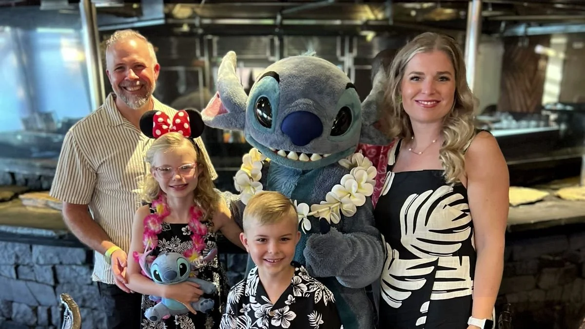 A family of four in blue and white hawaiian outfits pose with stitch at 'Ohana character breakfast at Disney World. Stitch is wearing a lei and you can see the Polynesian kitchen behind them.