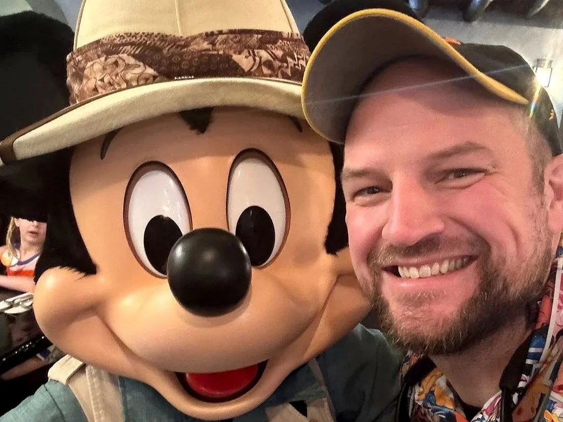 A man in a Disney character shirt poses with Mickey Mouse in a safari outfit. At Tusker House, a Disney World character dining restaurant at Animal Kingdom.