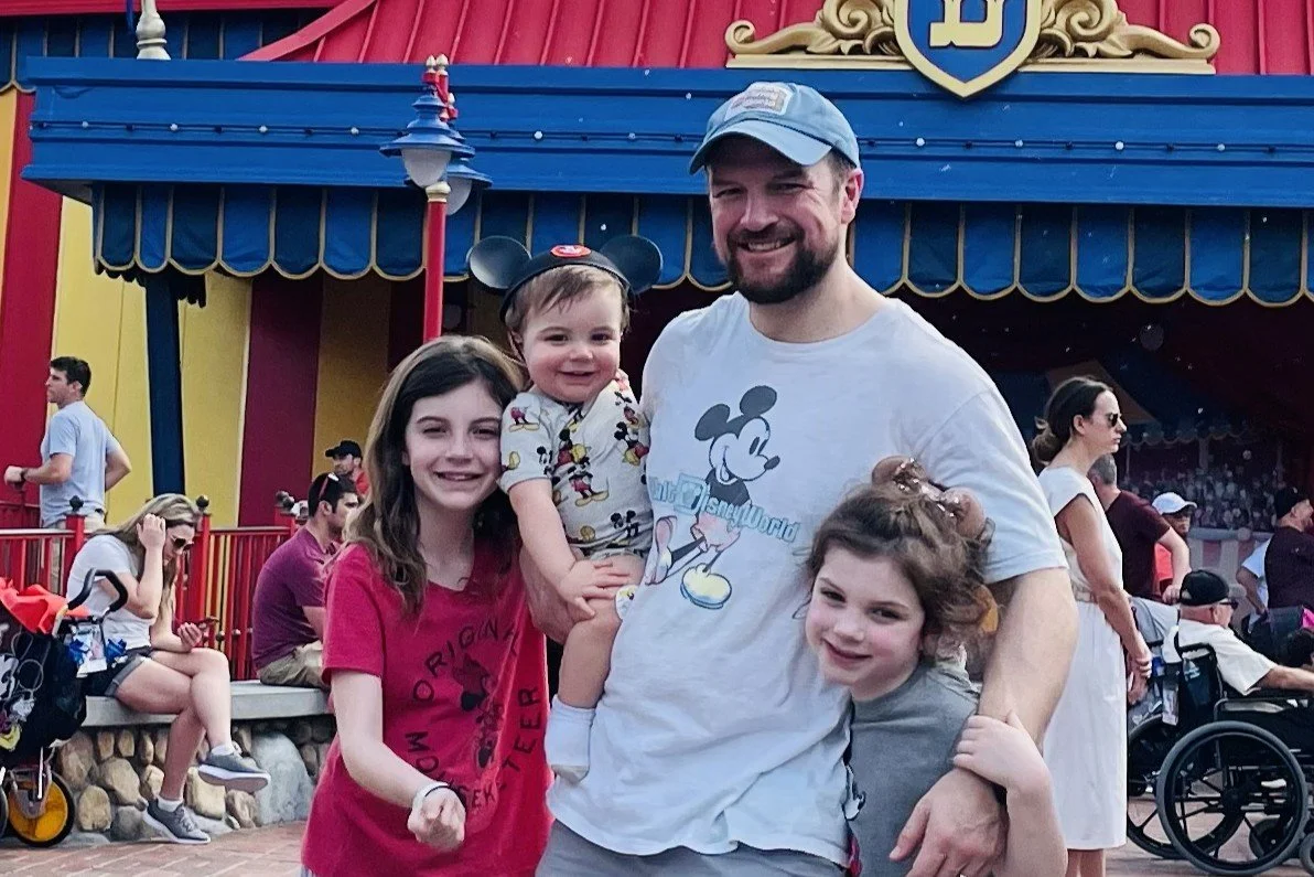 A family poses in Disney clothes, shorts, and t-shirts in front of the Dumbo ride. The baby has a Mickey ear hat on. They are in Magic Kingdom at Disney World in November