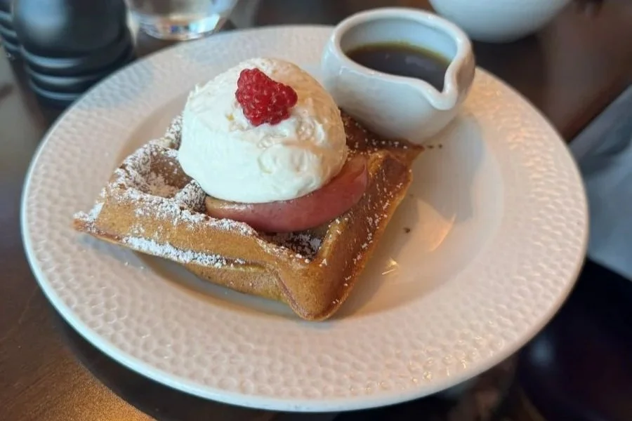 A photo of the sour cream waffle at Topolino's character breakfast. It's a square waffle with a peach and cream on top. Syrup in the background.