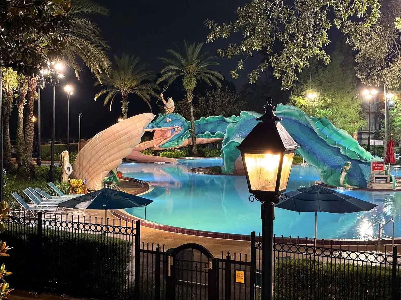 A night time picture of the Doubloon Lagoon pool at Disney's Port Orleans French Quarter at Disney World. You can see a curved pool with a giant sea monster structure winding through all of it. Poseidon rides the head, which is also a slide.