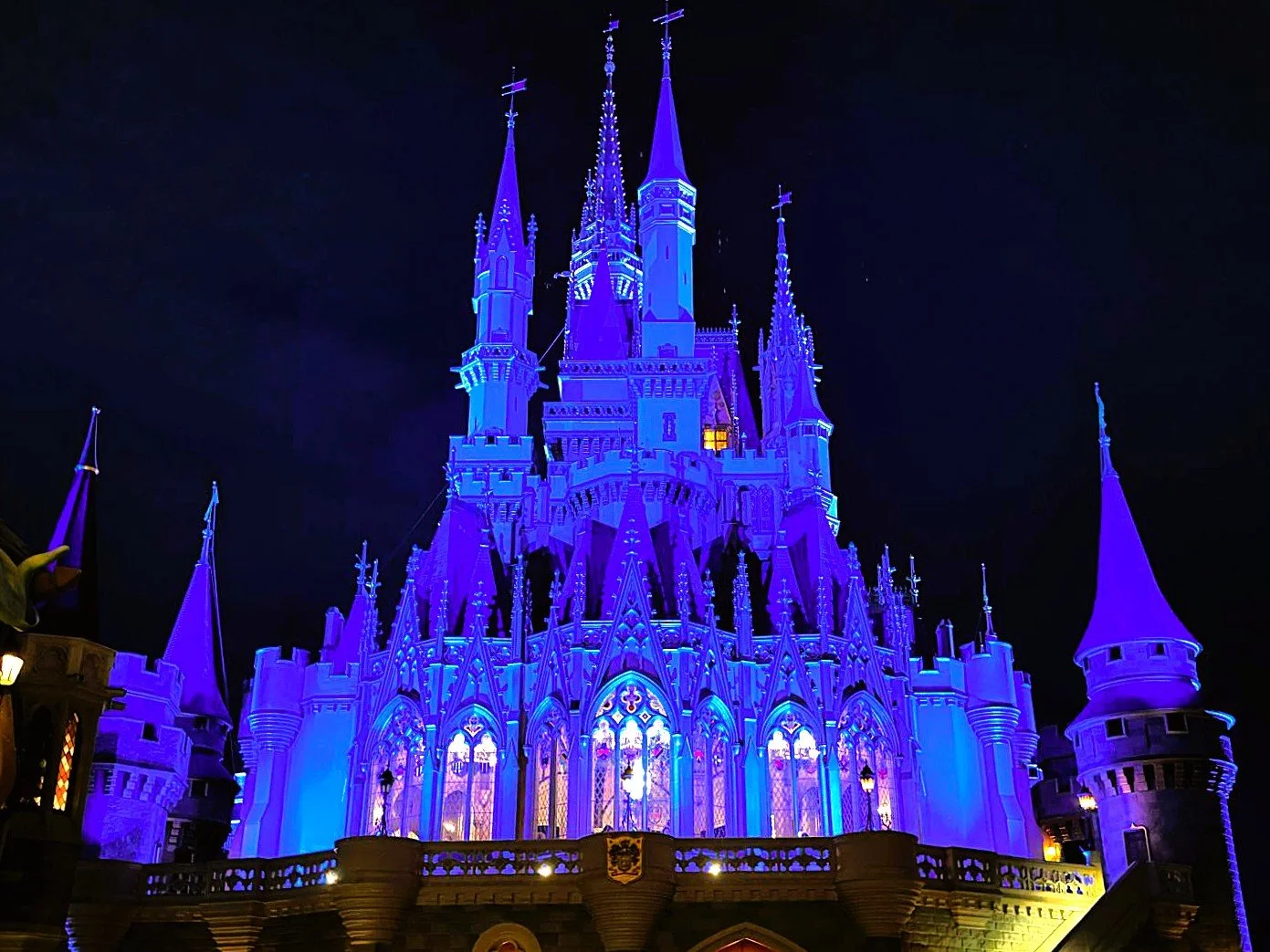 The back of Cinderella Castle, you can see several levels of spires on top of stone patio. The photo is at night and the lighting effects make the castle blue. Stained glass windows look into cinderella's royal table a character meal.