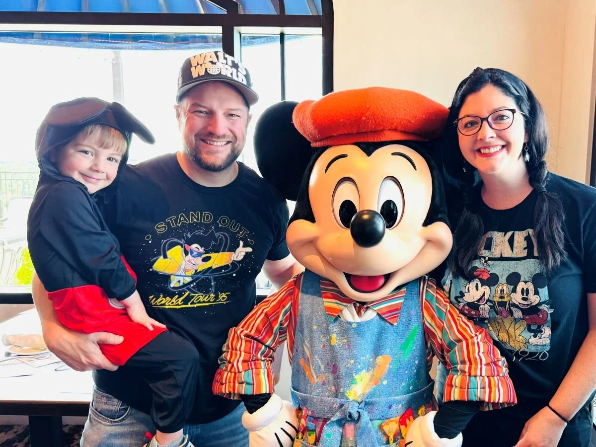 A father, son, and wife pose with Mickey. They are all in Mickey gear. Mickey is dressed as a painter at Topolino's character breakfast in Disney's Riviera Resort at Disney World.