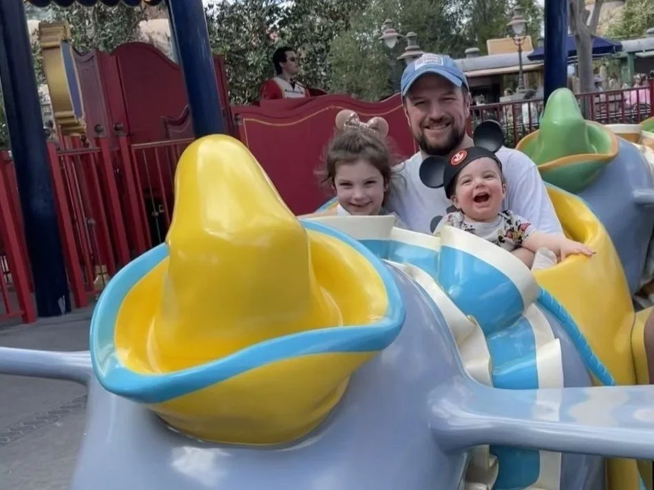 A 5 year old girl, an infant boy, and a dad smile at the camera while riding the Dumbo ride at Disney World's Magic Kingdom. The boy is wearing a crooked, classic mickey ears cap.
