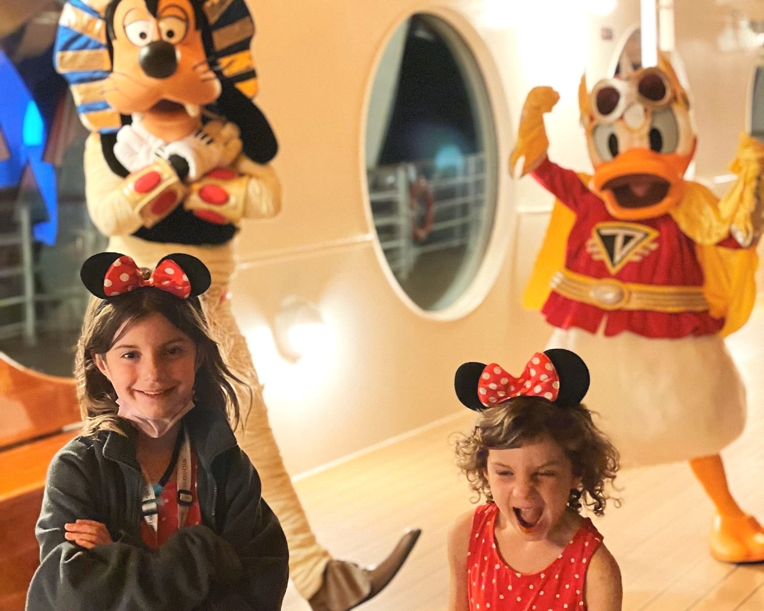 Two young girls with matching minnie mouse ears and dresses pose in front of goofy and donald dressed in halloween costumes on disney cruise for halloween on the high seas.