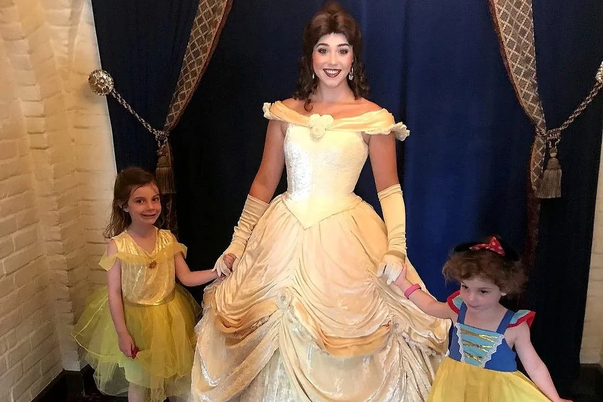 Two girls hold Belle's hand as they smile at the camera. Belle is wearing her formal yellow dress. At Akershus Royal Banquet Hall in EPCOT. A princess meal at Disney World.