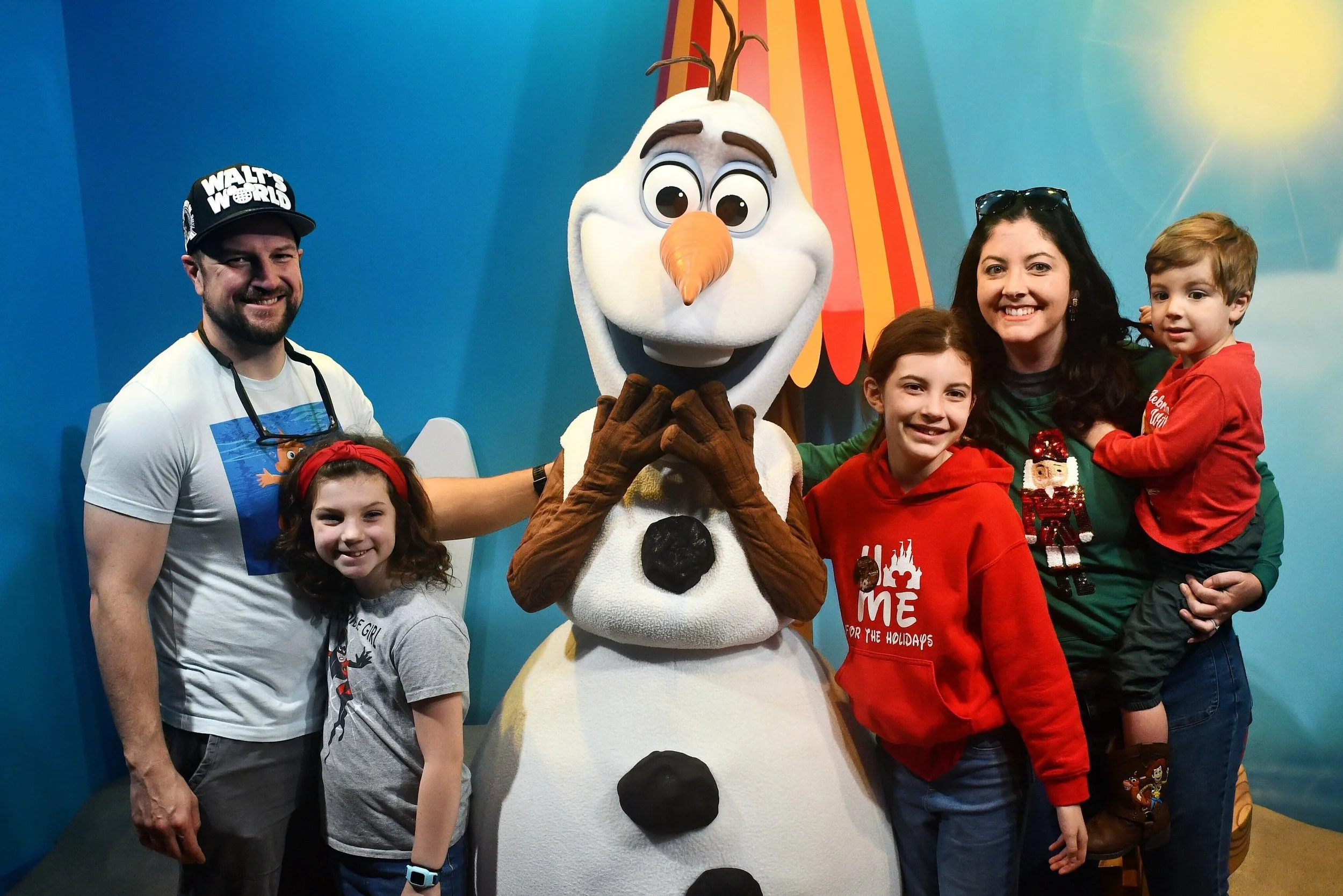 A family visits with Olaf. They are wearing an assortment of short sleeves, long sleeves, pants, and sweatshirts