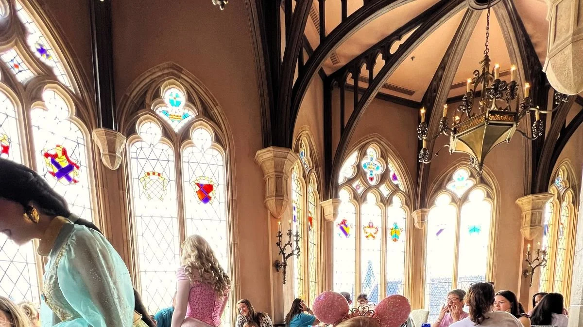 A photo of the dining room of Cinderella's Royal Table, a princess character meal at Disney World in the castle. You can see stained-glass windows with various crests, several princesses interacting, and lots of wood arches and buttresses.