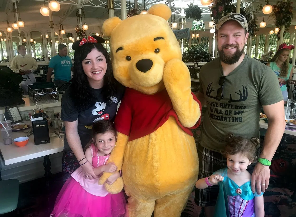 A family of four poses smiling with Winnie the Pooh, who is holding his hand to his cheek as a bashful pose. They are in the Crystal Palace character meal. You can see the many windows, ferns, skylight, and bulb lights behind them.