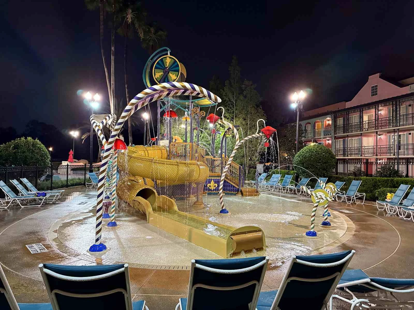 A nighttime picture of the Port Orleans French Quarter splash pad. You can see many whimsical spraying features, a large tunnel water slide, and Mardi gras themed splash feature at the top.