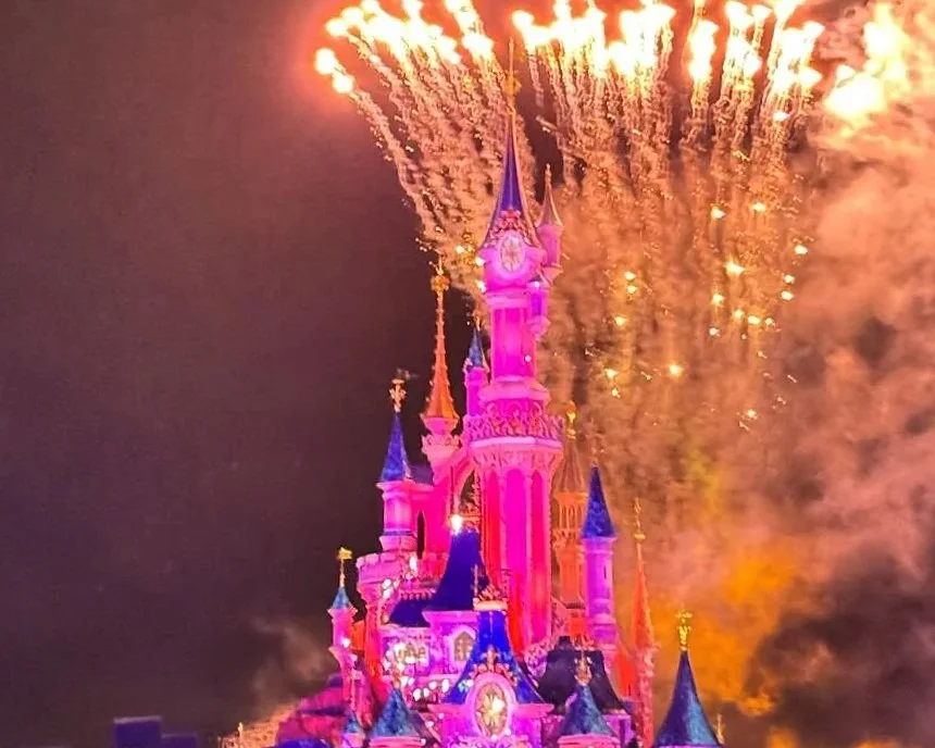 The Disneyland castle at night. The lights turn it pink and there are yellow fireworks exploding above it.