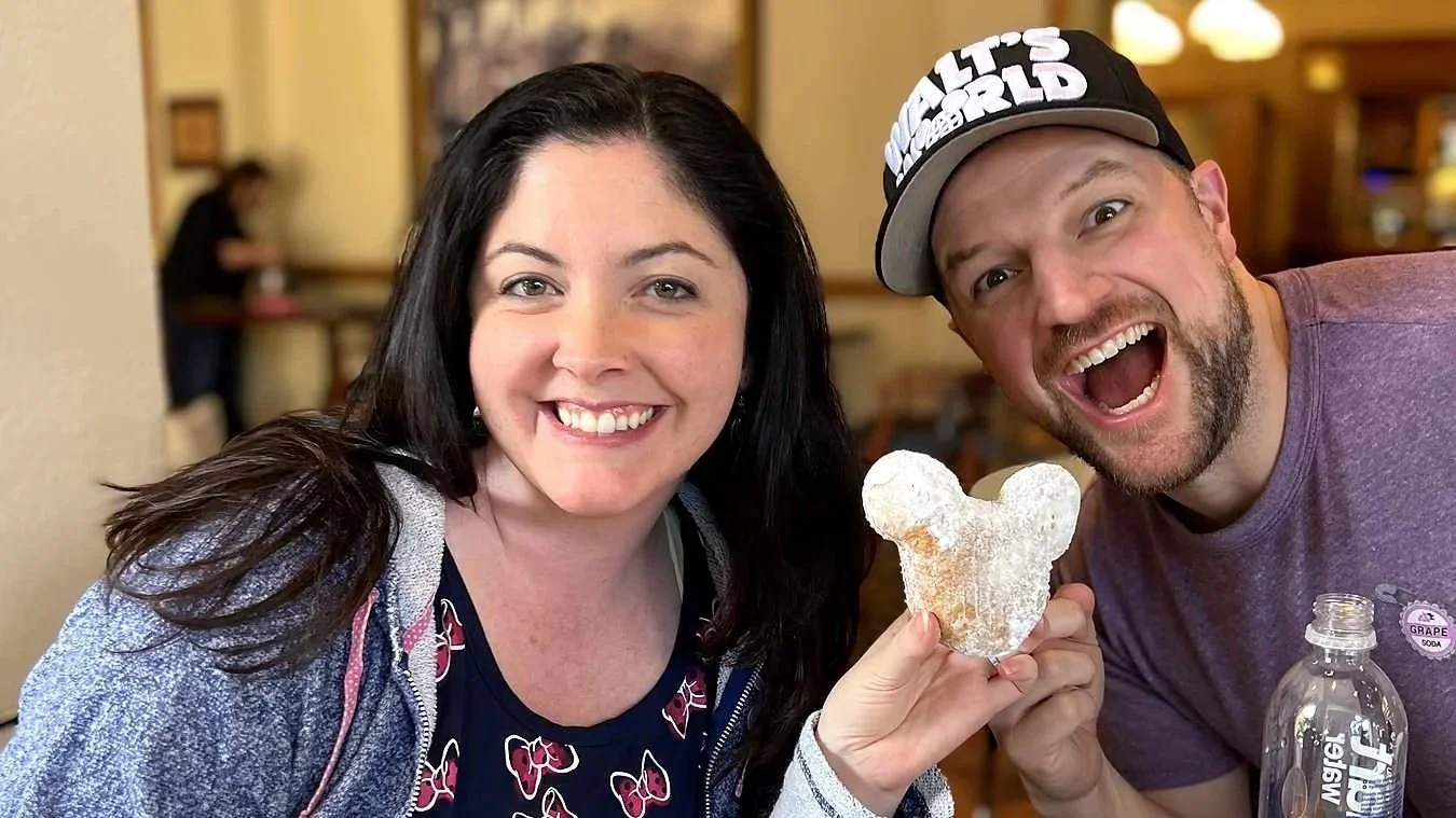 A couple smiles exaggeratedly while holding a mickey-shaped beignet at Port Orleans French Quarter, a Moderate Disney resort.