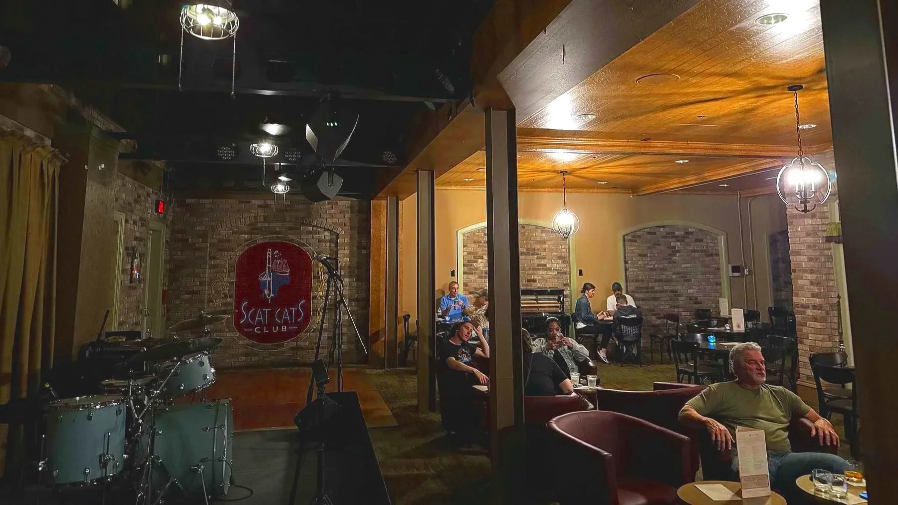 An image of Scat Cats Club Lounge at Disney's French Quarter resort. people sit in arm chairs with drinks on the right. the left has a sign, a stage, and a drum set visible. it all looks like a new orleans dive bar.