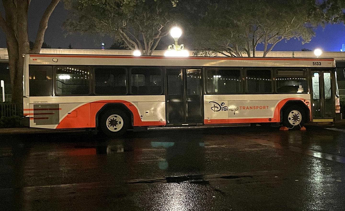 A photograph of a Disney bus outside the entrance of EPCOT at Disney World. The ground is wet from rain. It's nighttime.