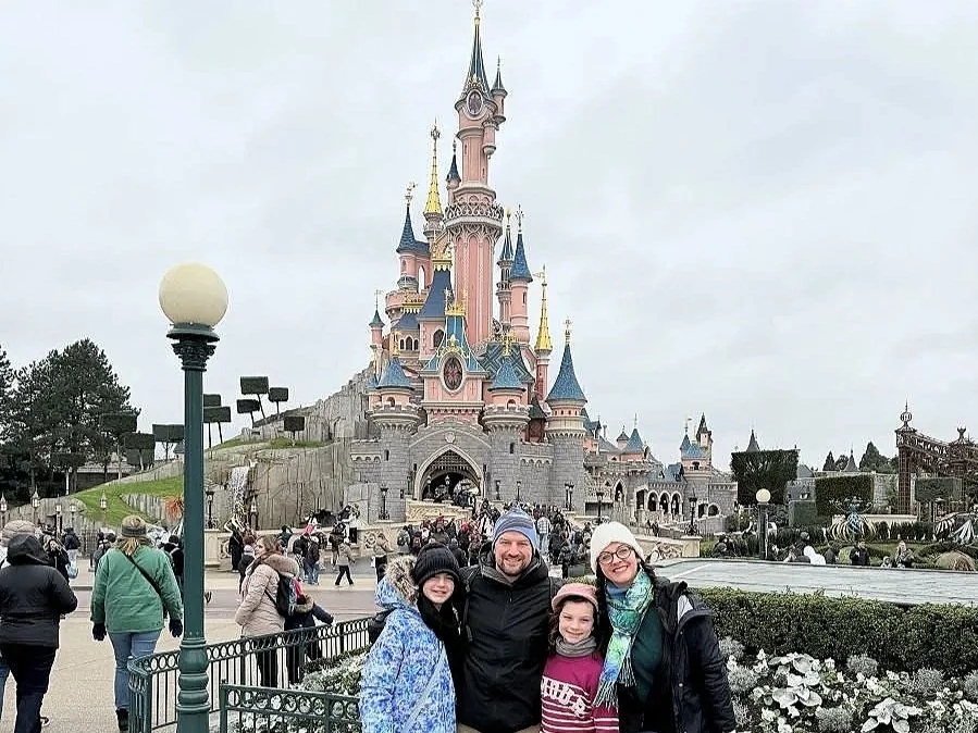 A family--dad, mom, two daughters-are standing in front of the disneyland paris castle. They are dressed for winter weather in France.