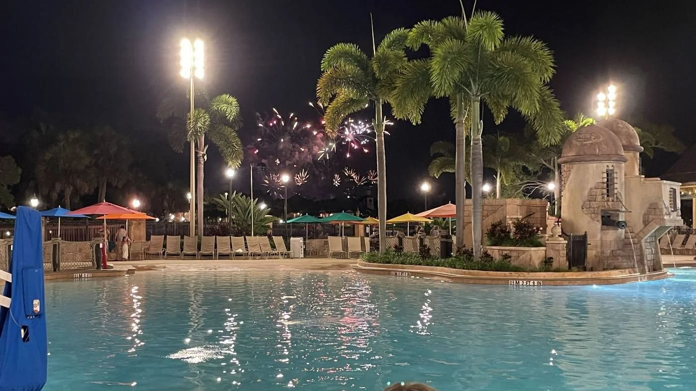 A view of the Caribbean Beach Resort pool at night time. You can see the EPCOT fireworks in the distance, just over the zero entry portion of the pool at this moderate Disney resort.