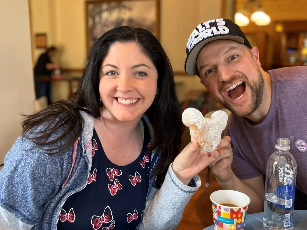 A photo of a couple holding up a Mickey-shaped beignet at Port Orleans French Quarter. It is covered in powdered sugar and there is a colorful coffee cup on the table. Both are wearing disney world themed shirts.