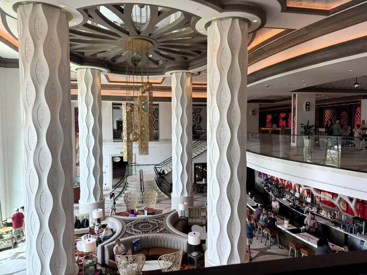 The lobby of the Gran Destino Tower, part of Coronado Springs, a moderate resort at Disney World. Towering white pillars drop down to the barcelona lounge, with lots of gold, white coral, and red light accents. Lots of irregular curves and marble.