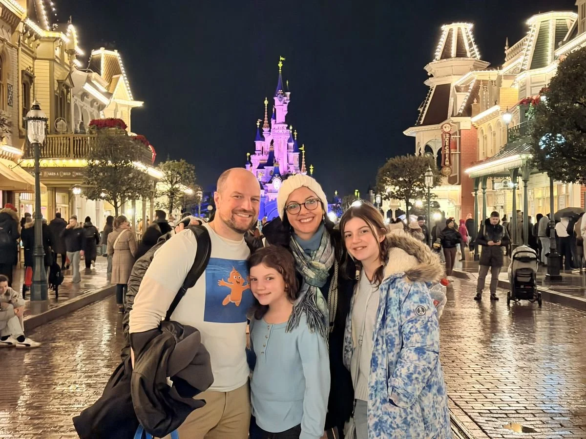 A family poses in winter clothes in front of the sleeping beauty castle in Disneyland Paris. Main street is lit on both sides.