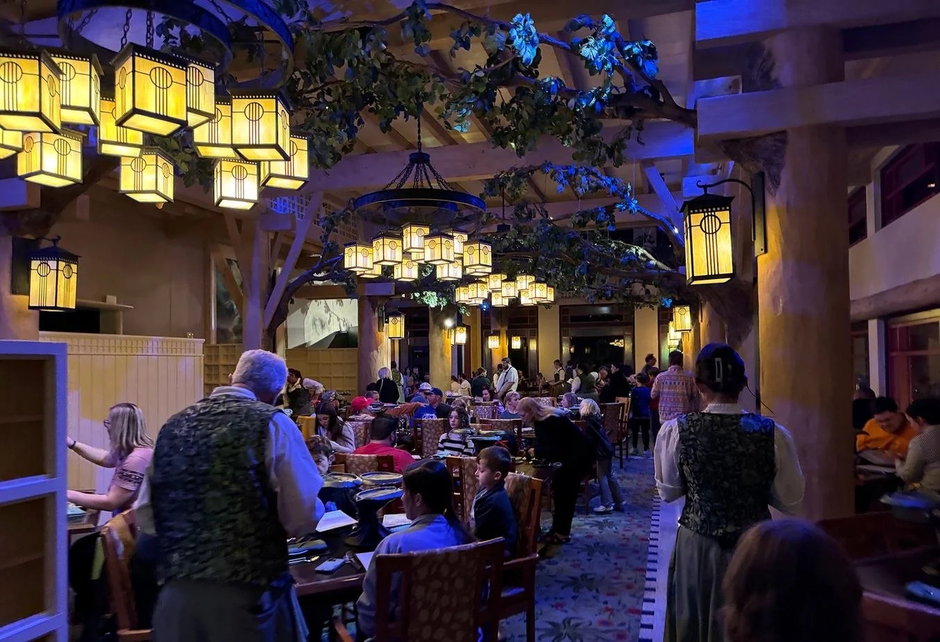 A view of the dining room at Story Book dining at Artist Point, a character dining experience with Snow White. It has enchanted forest vibe with a dark room, lantern chandeliers, and pillars shaped like trees with branches making a canopy.