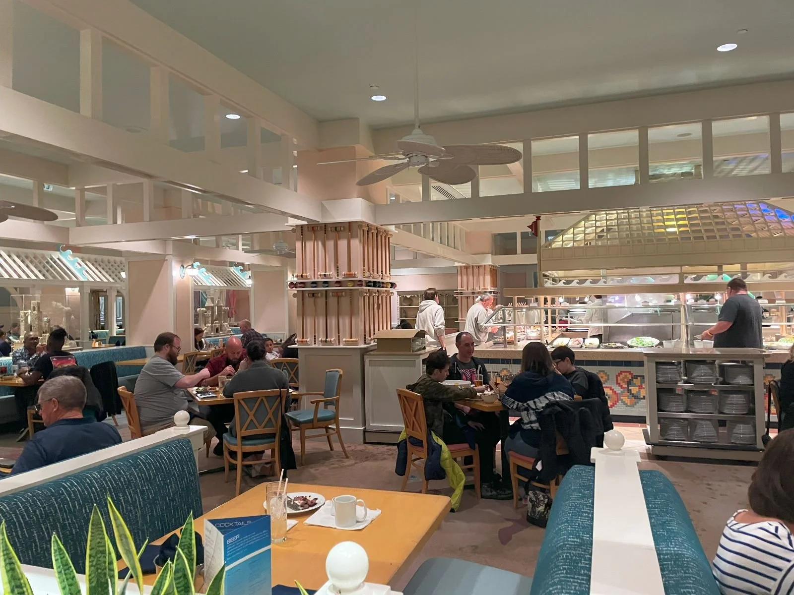 A photo of the interior of Cape May Cafe, a character dining experience inside Beach Club. You can see two rows of tables leading up to the buffet. It looks like a beach cottage with white walls, rectangular angles, and big bladed fans.