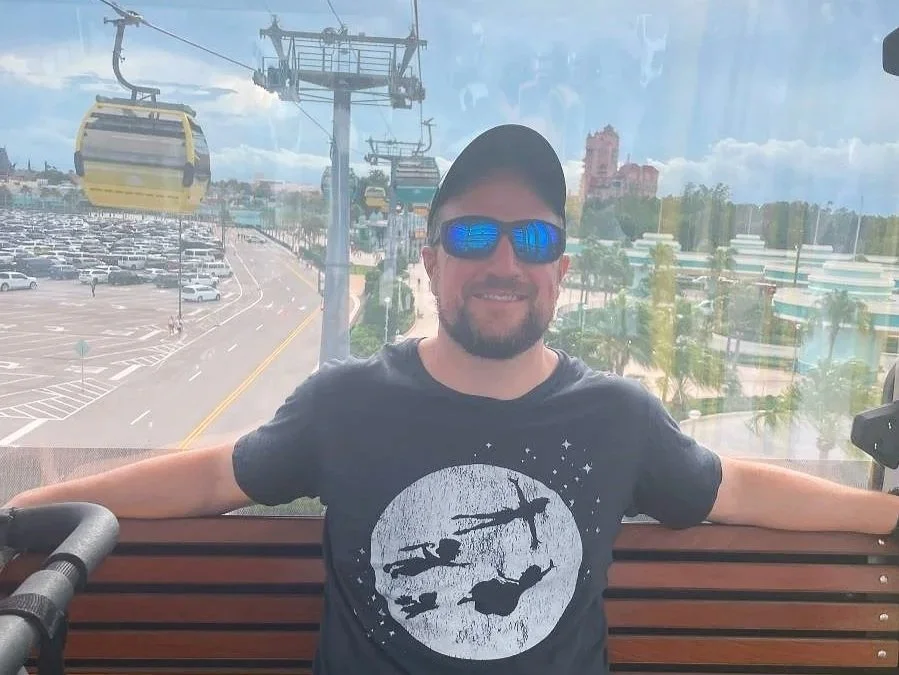 A man smiles with sunglasses and peter pan shirt while riding the skyliner. You can see a parking lot, other gondolas, and the tower of terror in the background at hollywood studios in disney world