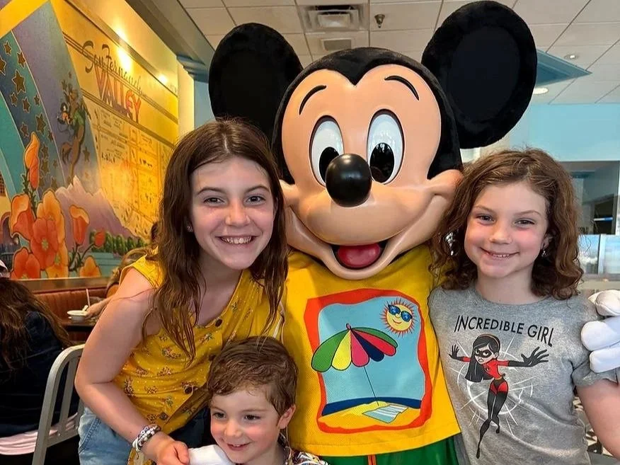 Three children smile as they pose with Mickey Mouse at Hollywood and Vine, a character dining restaurant in Hollywood Studios at Disney World. Mickey is wearing a t-shirt with a beach theme for their summer season outfit.