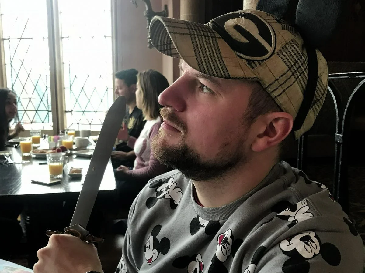A man with Mickey ears holds a plastic sword given as a souvenir at Cinderella's Royal Table, a Disney World character dining experience. He's holding it tight and looking into the distance as if to slay a dragon.