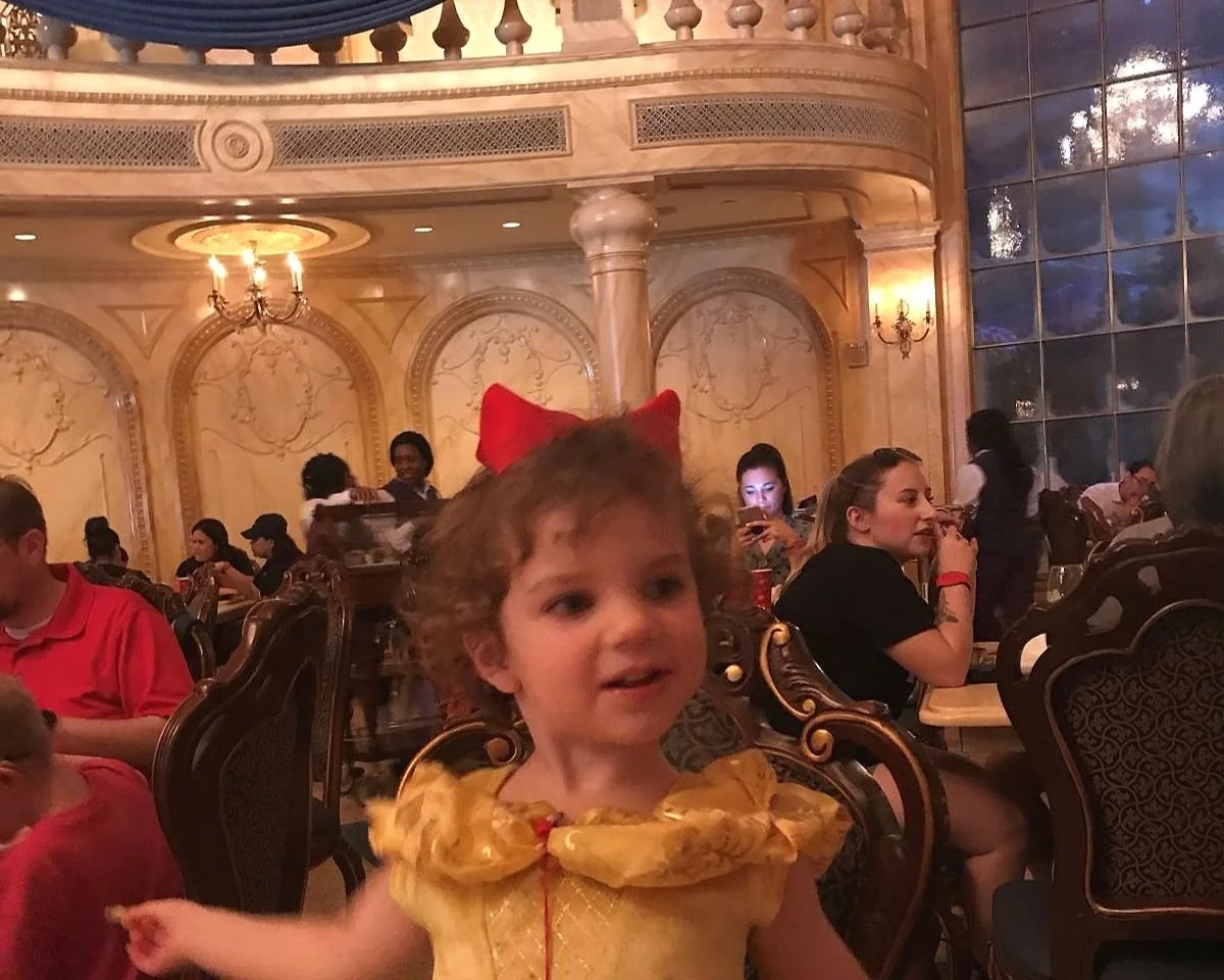 A girl in a Belle costume smiles as she eats food in the Be our guest restaurant, a Magic Kingdom restaurant themed to Beauty and the beast.