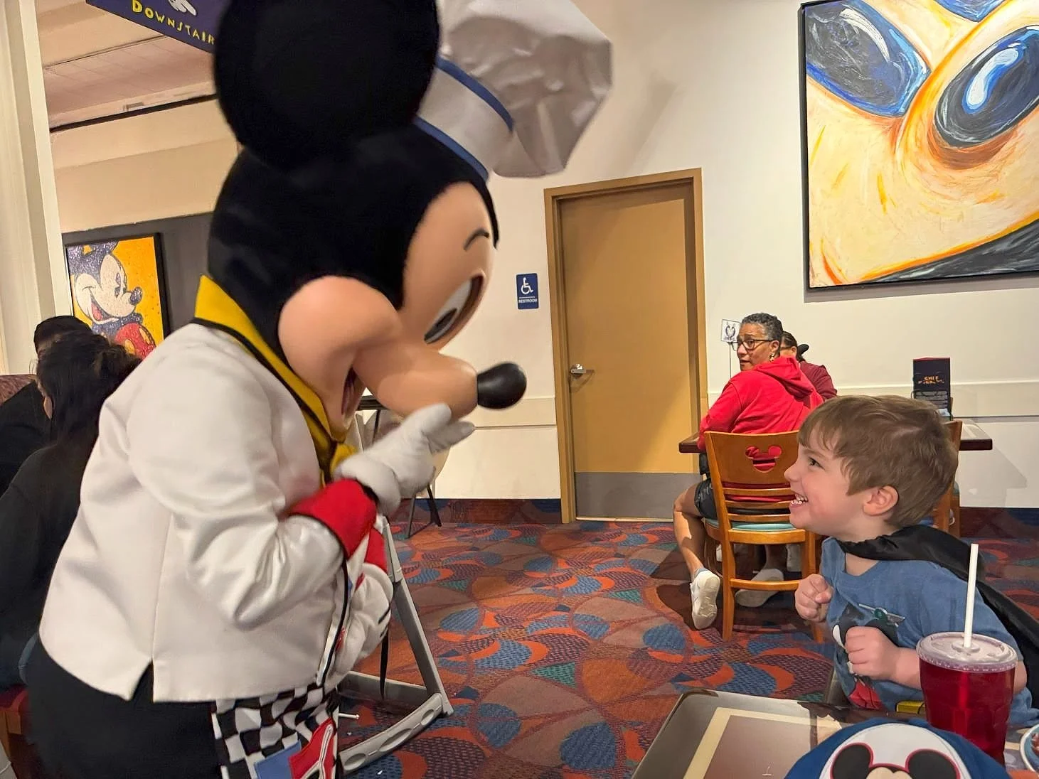 A toddler with a Mickey shirt and cape on smiles enthusiastically as Mickey laughs with his hand over his mouth. Mickey Mouse is dressed as a chef at Chef Mickey's character buffet dinner at Disney World.