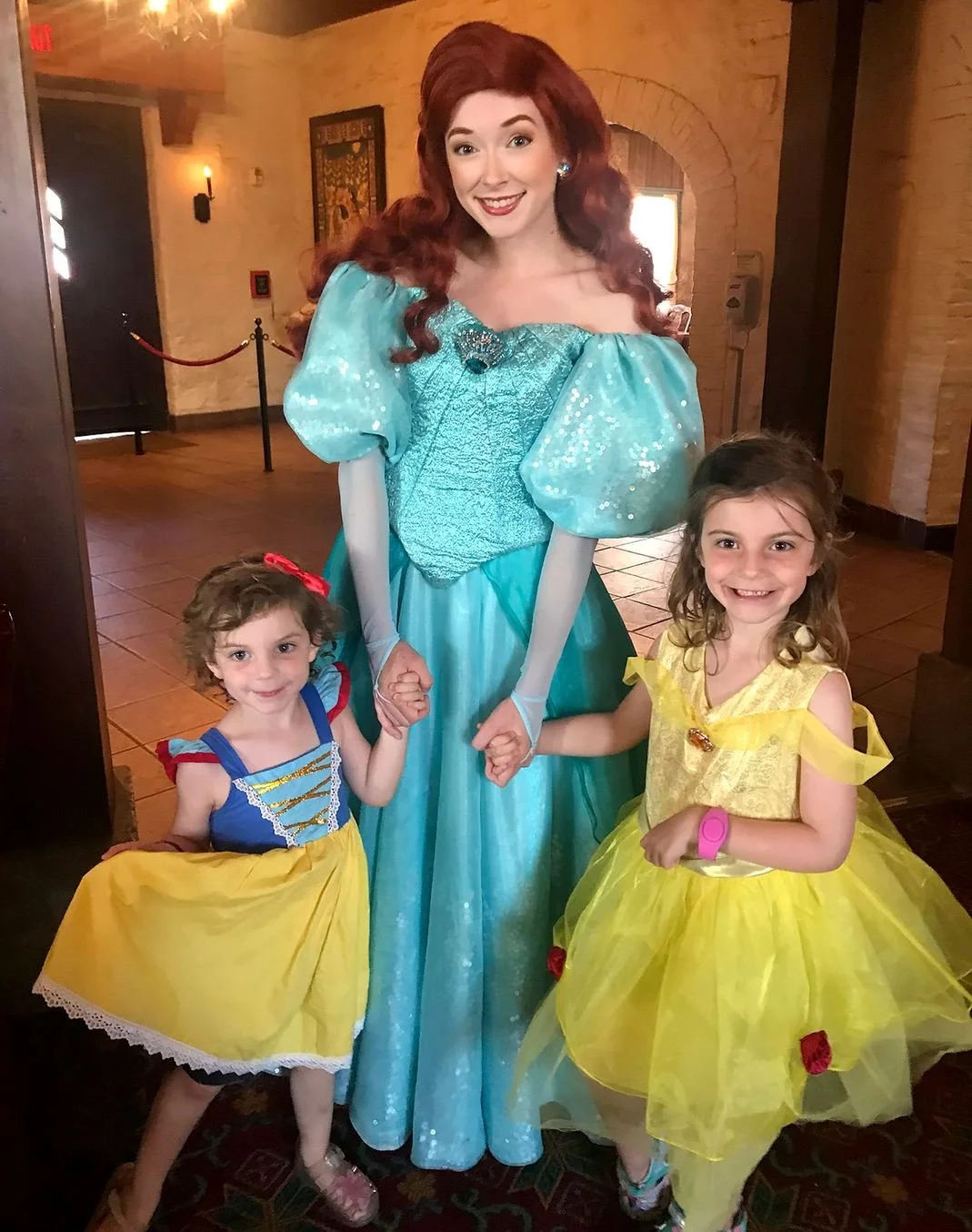 Ariel poses in a turquoise ball gown with two little girls also in princess costumes. Everyone is smiling, and the littlest is holding her dress to the side in a flourish. At Akershus Royal Banquet Hall in EPCOT, a Disney princess dining experience