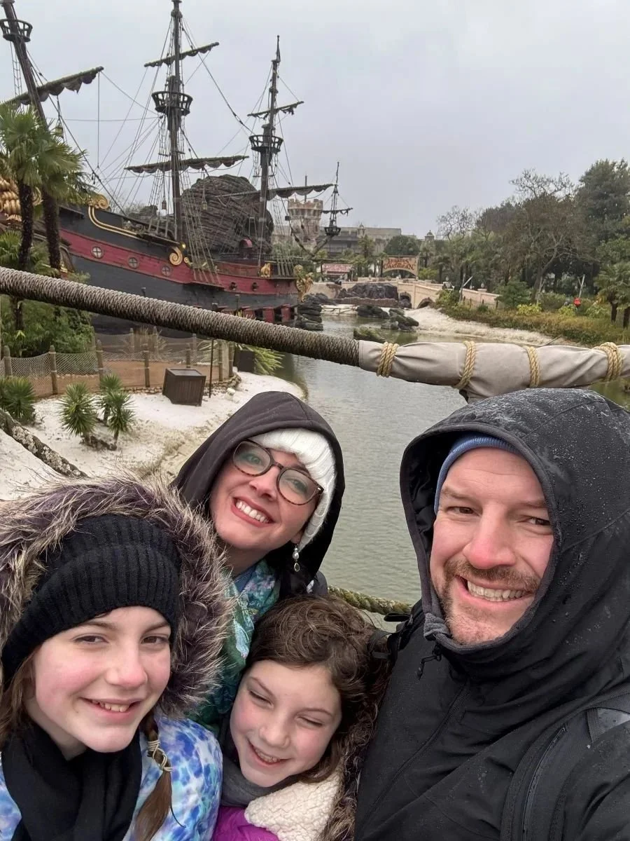 A family poses in front of adventureland in disneyland paris. There is snow on the dad's head.