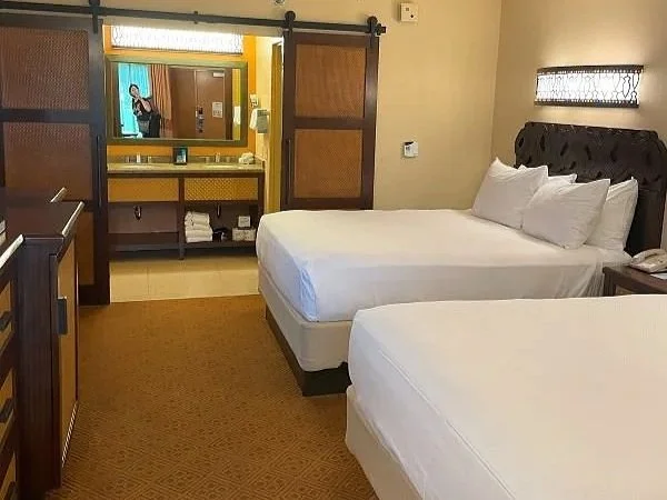 A double queen room at Caribbean Beach Resort. It has straw and wood sliding doors over the bathroom, and the headboard has dark wood. It has a tiki hut vibe. It's a moderate resort at Disney world.