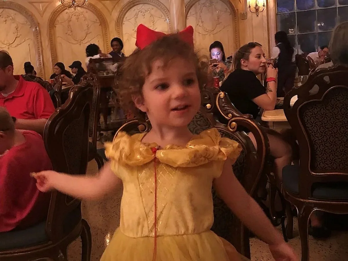 A little girl in a Belle dress eats a french fry in Be Our Guest restaurant in Magic Kingdom at Disney World. You can see the Beauty and the Beast ballroom theming behind her.