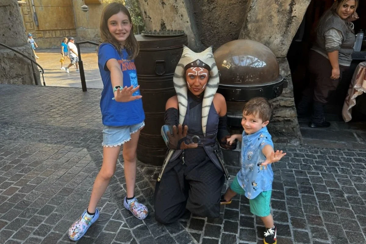 A sister and toddler brother pose with their hands outstretched with Ahsoka Tano from Star Wars in Disneyland in Star Wars land.