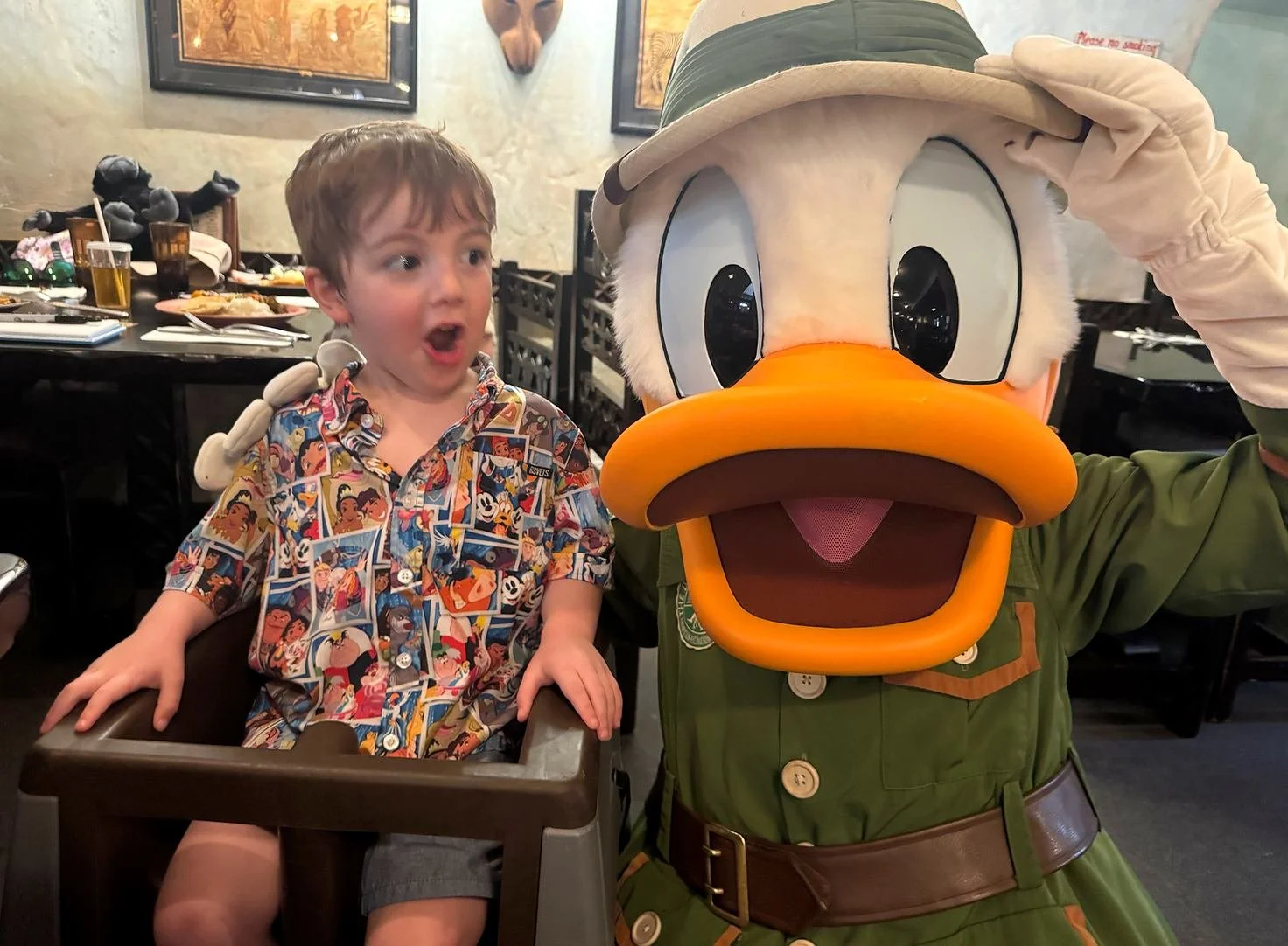Donald Duck in a green safari outfit poses on one knee with a toddler in a high chair. The boy is making a silly face. At Tusker House, an African-themed Disney World character dining restaurant at Animal Kingdom.