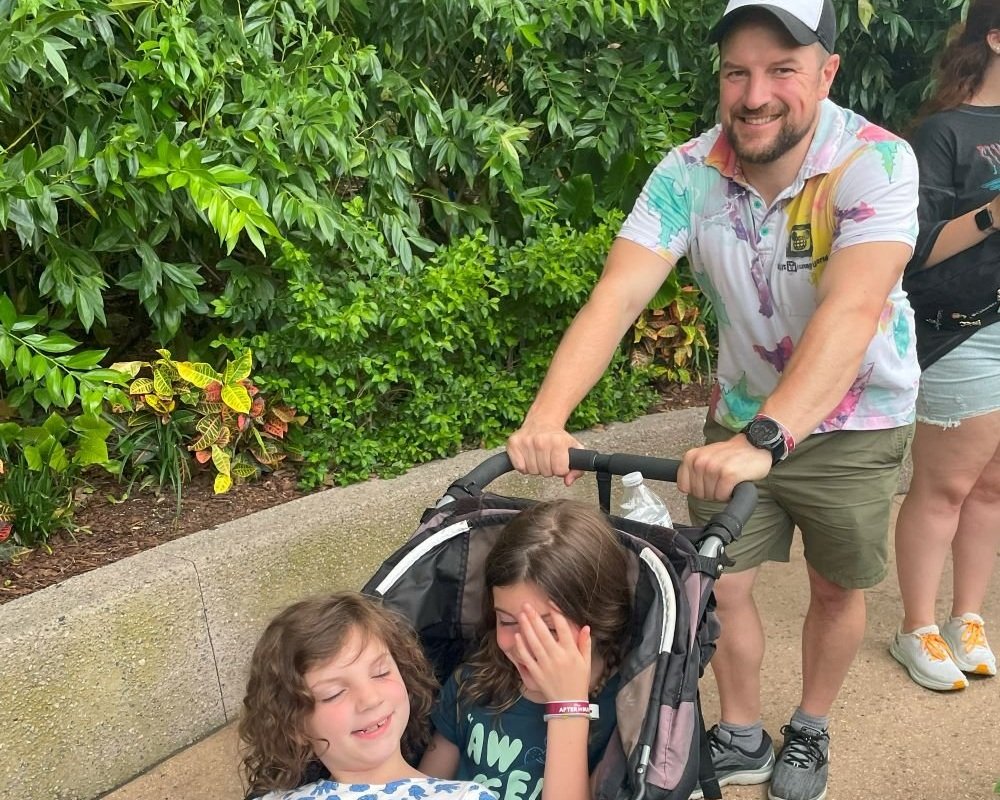 A dad in a Disney polo pushes his daughters in a BOB stroller during Disney After Hours EPCOT. Everyone is smiling awkwardly but excited to ride Frozen Ever After and Test Track.