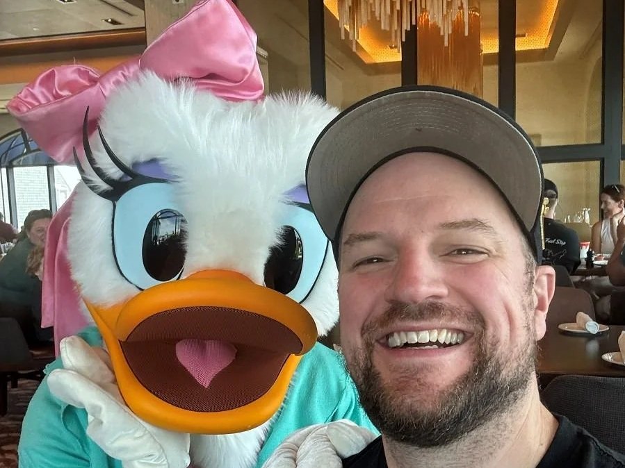 A man smiles in a hat while posing with Daisy Duck dressed as a ballerina. She has her hand on his shoulder and one under chin. At Topolino's character breakfast in the Riviera Resort at Disney World.