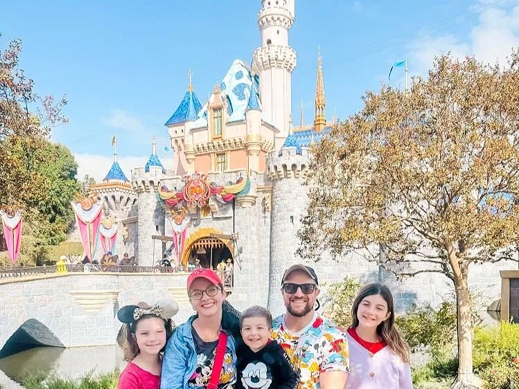 a family with one teenager, one elementary schooler, and one toddler pose on a family vacation in front of the disneyland castle in anaheim, california near los angeles. You can see the draw bridge and most of the turrets.